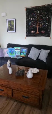 Sale Photo Thumbnail #37: Living room scene featuring a dark fabric sofa with matted artwork, a wooden coffee table displaying assorted pottery pieces, and a tree-of-life wall tapestry hanging above.