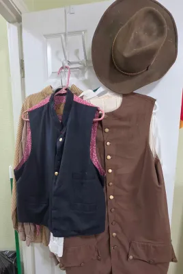 Sale Photo Thumbnail #71: Collection of Western and historical-style clothing, including a brown felt cowboy hat, buttoned waistcoats, and collared shirts hanging on a door rack.