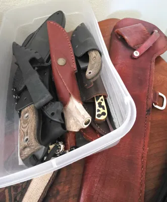 Sale Photo Thumbnail #9: Collection of fixed-blade hunting and tactical knives stored in a clear plastic container alongside leather and Kydex sheaths.