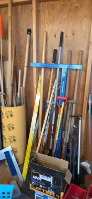 Sale Photo Thumbnail #33: Collection of long-handled yard tools, wooden dowels, drywall T-squares, and rulers leaning against an unfinished wall in a workshop.