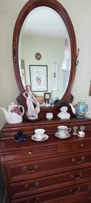 Sale Photo Thumbnail #80: Vintage dark wood dresser featuring a large oval swivel mirror, displayed with collectibles including decorative teapots, fine porcelain teacups with saucers, a figurine, and a china doll head.