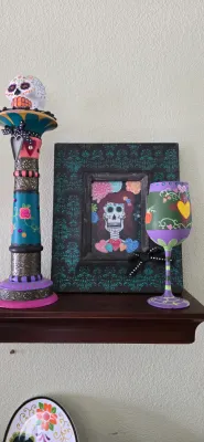 Sale Photo Thumbnail #102: Día de los Muertos decorative set featuring a colorful painted candlestick with sugar skull topper, a framed calavera print, and a hand-painted wine glass.