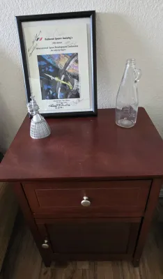 Sale Photo Thumbnail #47: Dark reddish-brown wood nightstand with a single top drawer and lower cabinet door, topped with space memorabilia including a framed 2001 International Space Development Conference poster, a small metallic rocket engine figurine, and a clear glass bottle.