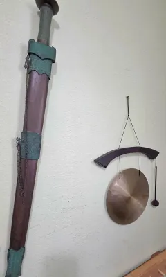 Sale Photo Thumbnail #60: Decorative Chinese-style Jian sword in a wooden scabbard with green fittings, mounted alongside a small hanging brass gong with a mallet.
