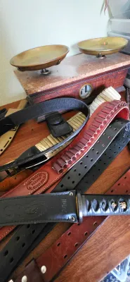 Sale Photo Thumbnail #12: Collection of gun belts, leather holsters, and tactical gear displayed alongside an antique double-pan balance scale on a marble base.