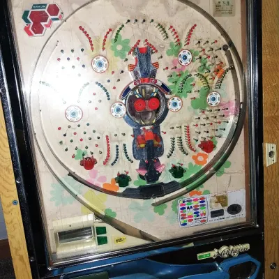 Sale Photo Thumbnail #4: A vintage mechanical Nishijin pachinko machine housed in a wooden frame. Features colorful playfield graphics, chrome accents, and mechanical internal components visible from the rear. Includes a small orange tray containing steel pachinko balls.