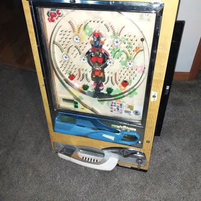Sale Photo Thumbnail #7: A vintage mechanical Nishijin pachinko machine housed in a wooden frame. Features colorful playfield graphics, chrome accents, and mechanical internal components visible from the rear. Includes a small orange tray containing steel pachinko balls.