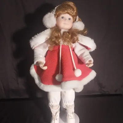 Sale Photo Thumbnail #429: Porcelain collector doll dressed in a winter ice skater outfit featuring a red coat trim with faux fur, matching earmuffs, white ice skates, and curly strawberry blonde hair. Hand-painted facial details with blue eyes. Comes complete with a metal display 