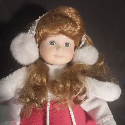 Sale Photo Thumbnail #430: Porcelain collector doll dressed in a winter ice skater outfit featuring a red coat trim with faux fur, matching earmuffs, white ice skates, and curly strawberry blonde hair. Hand-painted facial details with blue eyes. Comes complete with a metal display 