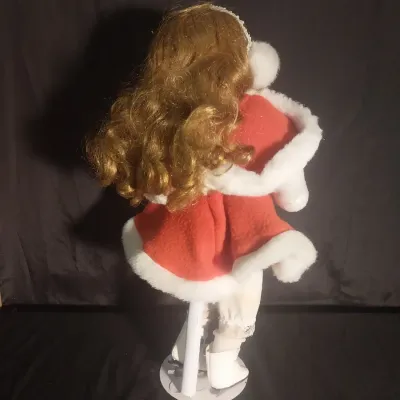 Sale Photo Thumbnail #431: Porcelain collector doll dressed in a winter ice skater outfit featuring a red coat trim with faux fur, matching earmuffs, white ice skates, and curly strawberry blonde hair. Hand-painted facial details with blue eyes. Comes complete with a metal display 