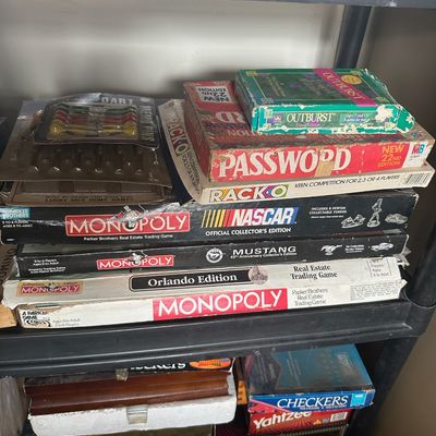 Collection of vintage board games stored on black plastic shelving, including multiple Monopoly editions, Password, Rack-O, Checkers, and...