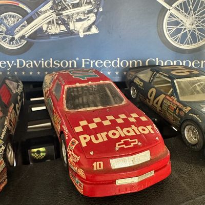 Vintage 1:24 scale NASCAR die-cast model cars featuring a red #10 Purolator Chevrolet Lumina, displayed in front of a Harley-Davidson...