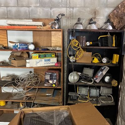 Collection of workshop lights, extension cords, clamp lamps, and portable halogen floodlights stored on wooden and metal shelving units.