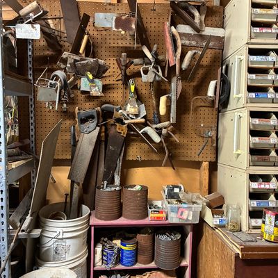 Collection of vintage hand tools including masonry trowels, hand saws, and scrapers arranged on a pegboard above storage shelving and...