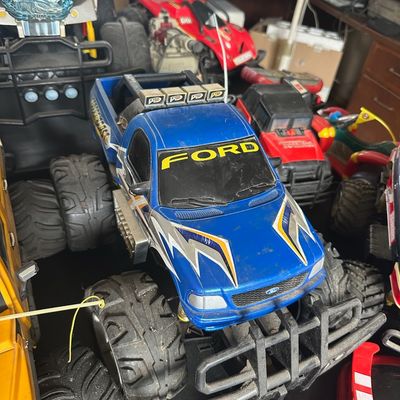 Collection of vintage toy monster trucks and remote-controlled vehicles, featuring a prominent blue Ford monster truck alongside red and...