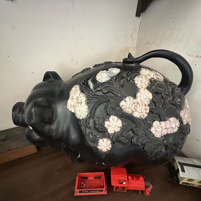 Vintage large black ceramic piggy bank featuring carved floral accents and a looped tail, shown alongside a small red toy train.