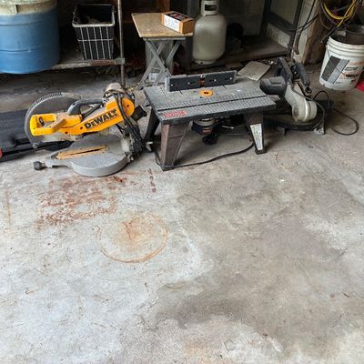 Collection of power tools including a yellow DeWalt miter saw, a Ryobi router table, and an additional miter saw sitting on a garage floor.