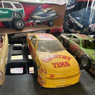 Vintage plastic and die-cast NASCAR toy race cars, featuring a Country Time Oldsmobile #68, alongside other collectible toy vehicles.