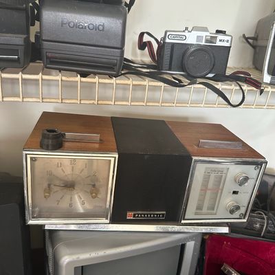 Vintage Panasonic woodgrain flip-clock radio sitting above a small portable CRT television, alongside Polaroid instant cameras and a...