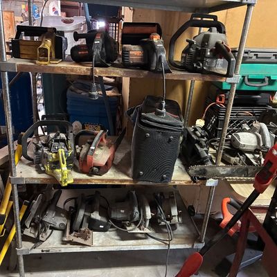 Wire shelving unit filled with vintage power tools, including chainsaws, circular saws, and planers showing moderate use and wear.