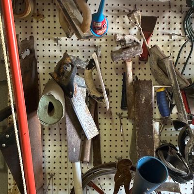 Assorted vintage hand tools, including masonry floats, handsaws, hammers, and funnels, hanging on a white pegboard.