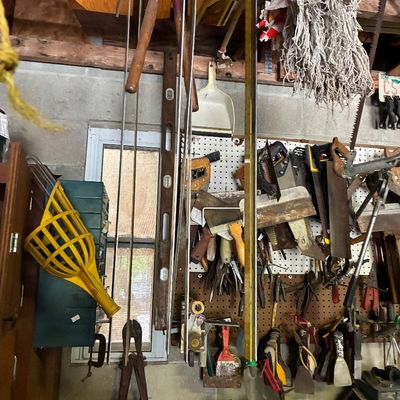 Collection of assortment garage tools, including handsaws, loppers, bubble levels, and fruit pickers, hanging from a pegboard wall and...