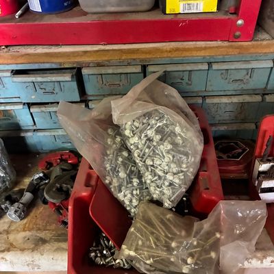 Assorted workshop hardware, including bagged roofing screws, loose bolts in a red plastic organizer bin, and pipe cutters on a workbench.