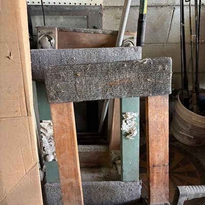 Pair of homemade wooden boat stands lined with grey carpet, stored in a garage alongside fishing equipment.