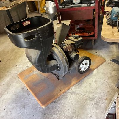 Gas-powered wood chipper and shredder mounted on a wheeled base, resting on a wooden board in a workshop setting.