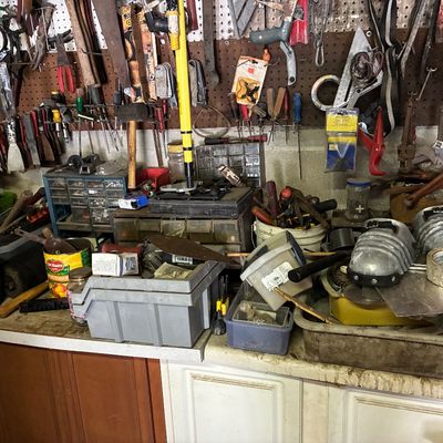 Cluttered workshop bench and pegboard filled with vintage hand tools, hardware organizers, knee pads, and various hardware supplies.