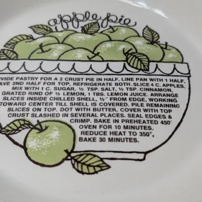 Sale Photo Thumbnail #149: The vintage 1982 Royal China by Jeannette 11" Apple Pie Recipe Plate is a highly collectible, oven-to-table ceramic dish. Featuring a fluted edge and a distinct green and brown apple motif, it lists the exact ingredient amounts and baking instructions dir