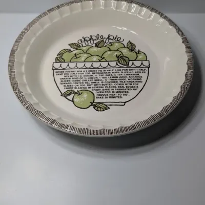 Sale Photo Thumbnail #148: The vintage 1982 Royal China by Jeannette 11" Apple Pie Recipe Plate is a highly collectible, oven-to-table ceramic dish. Featuring a fluted edge and a distinct green and brown apple motif, it lists the exact ingredient amounts and baking instructions dir