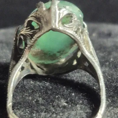 Sale Photo Thumbnail #326: Vintage filigree-style ring featuring an ornate silver mounting with scrollwork and milgrain detailing, set with a large oval green cabochon stone held by claw prongs. The band is split-shank style with decorative open metalwork on the shoulders. Pair of 