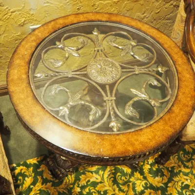 Sale Photo Thumbnail #147: Decorative round side table featuring a clear glass inset displaying elaborate metal scrollwork beneath, surrounded by a carved wood border. Supported by a substantial carved pedestal and sculptural base. 27.5" round by 28 h