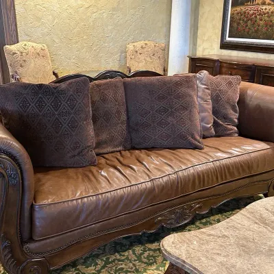 Sale Photo Thumbnail #108: Large traditional sofa upholstered in warm brown leather with oversized rolled arms, nailhead trim, carved wood base, and decorative scroll detailing. Includes coordinating oversized accent pillows and attractive aged character. 91w*38 deep* 41h
