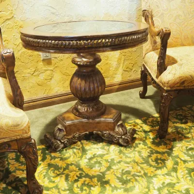 Sale Photo Thumbnail #146: Decorative round side table featuring a clear glass inset displaying elaborate metal scrollwork beneath, surrounded by a carved wood border. Supported by a substantial carved pedestal and sculptural base. 27.5" round by 28 h