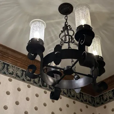Sale Photo Thumbnail #166: Traditional four-light chandelier featuring a circular wrought-iron frame, heavy scrollwork, and cylindrical crackle-glass hurricane shades. Strong Spanish Revival / Old World styling with dramatic light patterns when illuminated. Buyer responsible for re