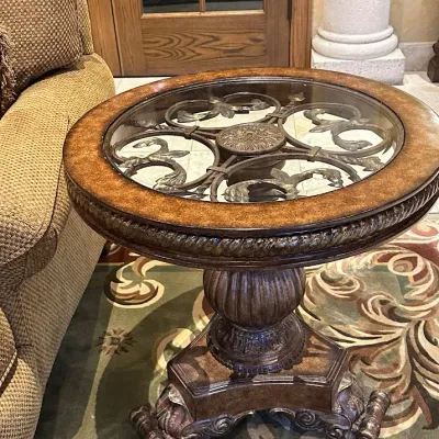 Sale Photo Thumbnail #145: Decorative round side table featuring a clear glass inset displaying elaborate metal scrollwork beneath, surrounded by a carved wood border. Supported by a substantial carved pedestal and sculptural base. 27.5" round by 28 h