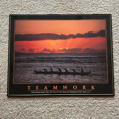 Sale Photo Thumbnail #114: This motivational poster titled 'TEAMWORK' showcases a striking silhouette of six individuals paddling a canoe against a vivid orange and pink sunset over calm waters. The black and gold bordered frame complements the warm hues of the sky, creating a visu