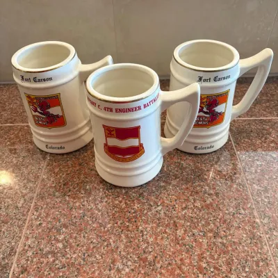Sale Photo Thumbnail #299: This set includes three ceramic beer steins, each standing 6 inches tall with a diameter of 3.5 inches. The steins feature colorful historic insignias from Fort Carson, Colorado, including unit crests and emblems for Delta Demons and Company C, 4th Engine