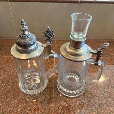 Sale Photo Thumbnail #296: This German style beer stein set features two unique glass steins adorned with intricately detailed pewter lids and lion-themed thumb lifts. The set includes a 9-inch tall stein with an ornate lion thumb lift, a 7.5-inch stein without a shot glass, and a 