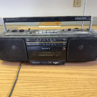 Sale Photo Thumbnail #328: This vintage Sony CFD-S12OS boombox radio features a classic black design with a sturdy plastic casing. It includes dual built-in speakers and a Mega Bass feature enhancing audio quality. The device supports cassette tape playback and radio tuning with an
