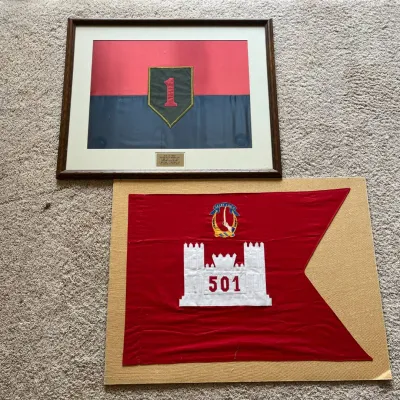 Sale Photo Thumbnail #57: The first is a framed red-and-blue textile display featuring the distinctive “Big Red One” 1st Infantry Division insignia. A mounted plaque identifies officer and documents his service as Secretary of the General Staff from June 8, 1987–July 7, 1988
