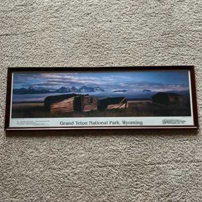 Sale Photo Thumbnail #113: This framed panoramic photograph by Leo L. Larson captures the majestic landscape of Grand Teton National Park in Wyoming, featuring the iconic Pfeiffer Homestead and Teton Range in the background. The image is printed with a rich color palette that highl