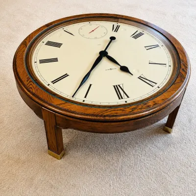 Sale Photo Thumbnail #185: This unique coffee table features a working Howard Miller clock integrated into a solid oak frame. The table stands 16 inches tall and has a 41.5-inch round top, showcasing an elegant clock face with Roman numerals and a small seconds dial. The rich oak w