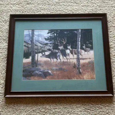 Sale Photo Thumbnail #81: This beautifully framed wildlife art print showcases a serene scene of five deer standing in a grassy meadow with a backdrop of trees and distant mountains. The print is set within a classic dark wood frame that features subtle carved details and a rich b