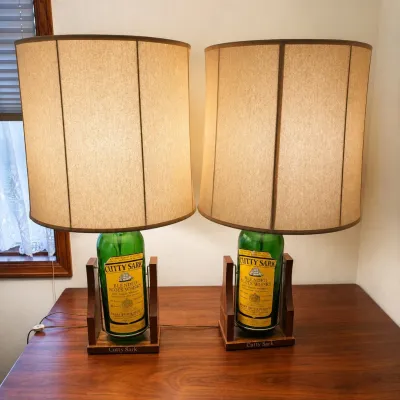 Sale Photo Thumbnail #10: This unique set features two vintage Cutty Sark Blended Scots Whisky bottles repurposed as stylish table lamps. Each lamp includes a 17-inch tall bottle with authentic yellow and green Cutty Sark labels, mounted on a custom wooden base engraved with 'Cutt