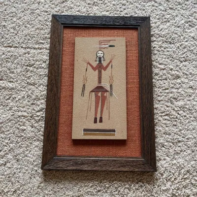 Sale Photo Thumbnail #95: Vintage framed sand art depicting a stylized ceremonial figure in traditional Southwestern-inspired colors and geometric forms. The image is created on a textured sand-like surface in shades of rust, brown, black and white and is mounted over a woven rust