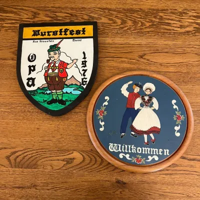 Sale Photo Thumbnail #330: This set includes a vintage Wurstfest 1976 shield featuring a colorful illustration of an Opa in traditional attire, marked with 'New Braunfels, Texas,' and a round wooden Willkommen plaque showcasing a lively folk dance couple against a rich blue backgro