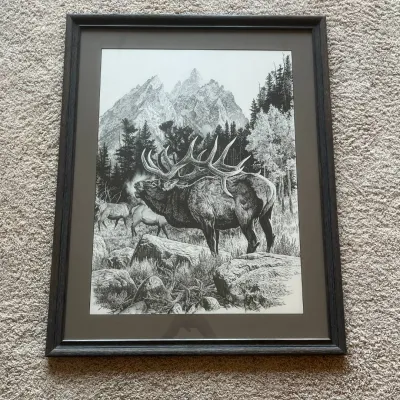 Sale Photo Thumbnail #80: This framed black and white wildlife print depicts a majestic elk with large antlers standing in a forest clearing with additional elk and towering mountain peaks in the background. The artwork showcases detailed stippling technique and is signed by the a